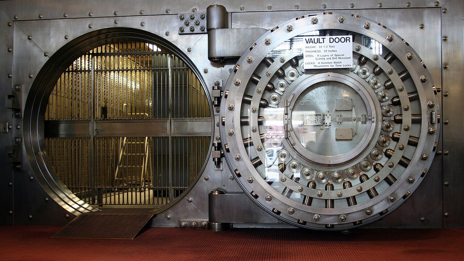 A massive round steel bank vault door with brass details in a dark, blue-lit chamber